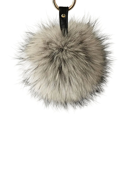 A fluffy REAL-fur pom-pom keychain in shades of gray, beige, and white, featuring a black leather strap with a gold rivet and a gold split ring at the top, set against a plain light gray background.