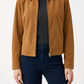 A woman wearing a cropped tan suede jacket over a black top and dark blue jeans, shown from the neck down against a plain white studio background.